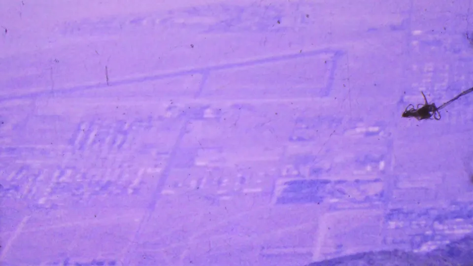 1964 8mm aerial view over Palm Springs with housing rows, open desert plots, a hillside edge, and a prominent dark dust or debris mark near the center.