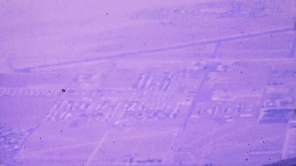 Soft purple-shifted 1964 aerial view of Palm Springs, showing faint roads, housing rows, open desert land, and blurred built areas below the tramway vantage.