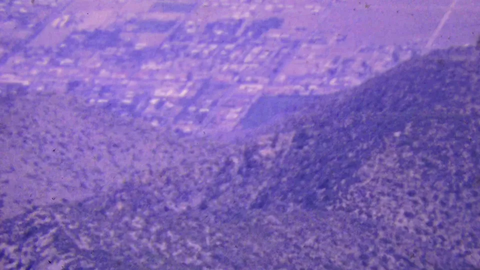 The frame looks down over a Palm Springs road grid, small structures, and adjacent desert hillside terrain in grainy purple-shifted 8mm footage.
