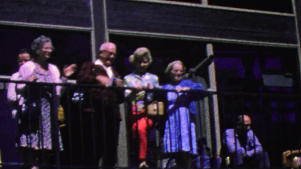 Older tourists remain gathered on the Palm Springs tramway balcony, framed by dark rails and shaded exterior structure as one man waves toward the camera.