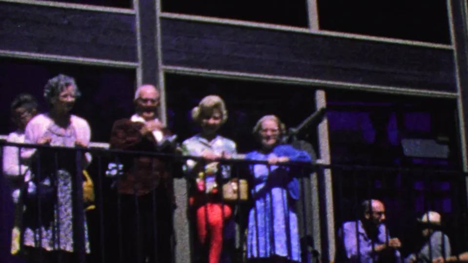 The 1964 home-movie frame shows older tourists on a Palm Springs tramway balcony, with dark rails, shaded structural posts, and colorful period clothing visible.