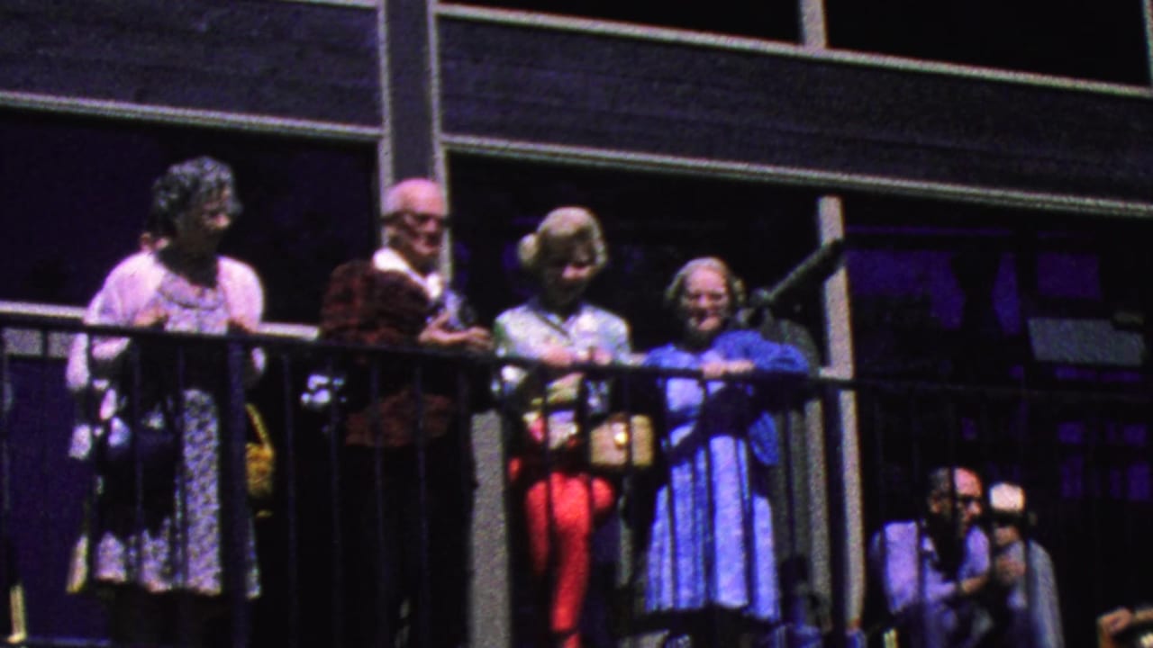 Elderly tourists stand behind a black balcony railing outside a glass-fronted building.