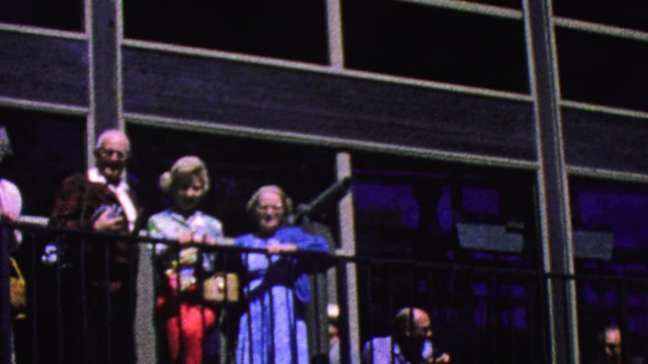 A group of older visitors leans on a balcony railing near large windows.
