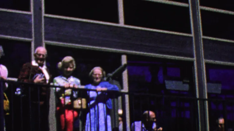Older visitors in 1960s clothing lean and stand along the Palm Springs tramway balcony with large framed windows behind them.