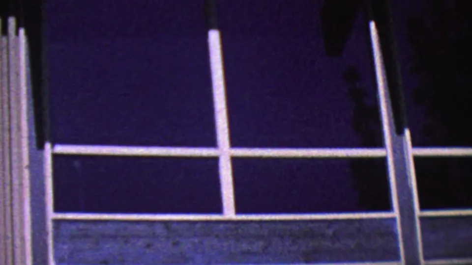 1964 Palm Springs home-movie frame of a large white-mullioned window grid beneath a dark overhang, with purple-blue sky and faint tree silhouettes.