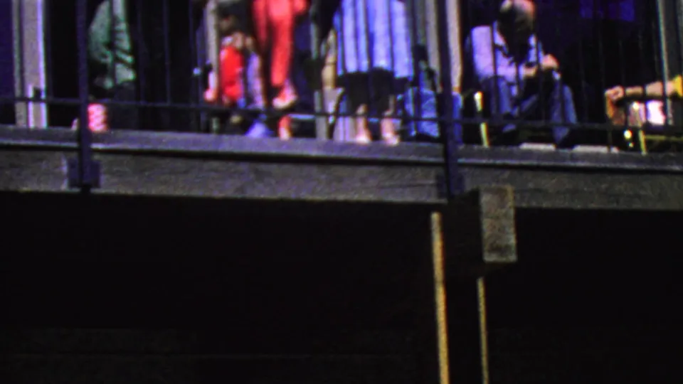 A dark, grainy 8mm frame from the Palm Springs Aerial Tramway visit shows visitors on an upper balcony with railings, vertical siding, and shadowed building recesses below.