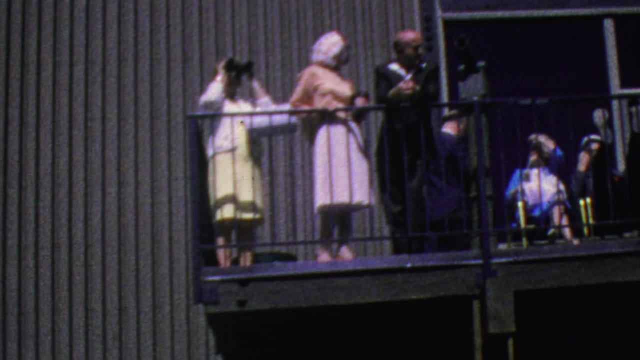 1964 Palm Springs Aerial Tramway visitors stand on an outdoor balcony, including a woman with binoculars, a woman in a pink dress, and a man in a dark suit.