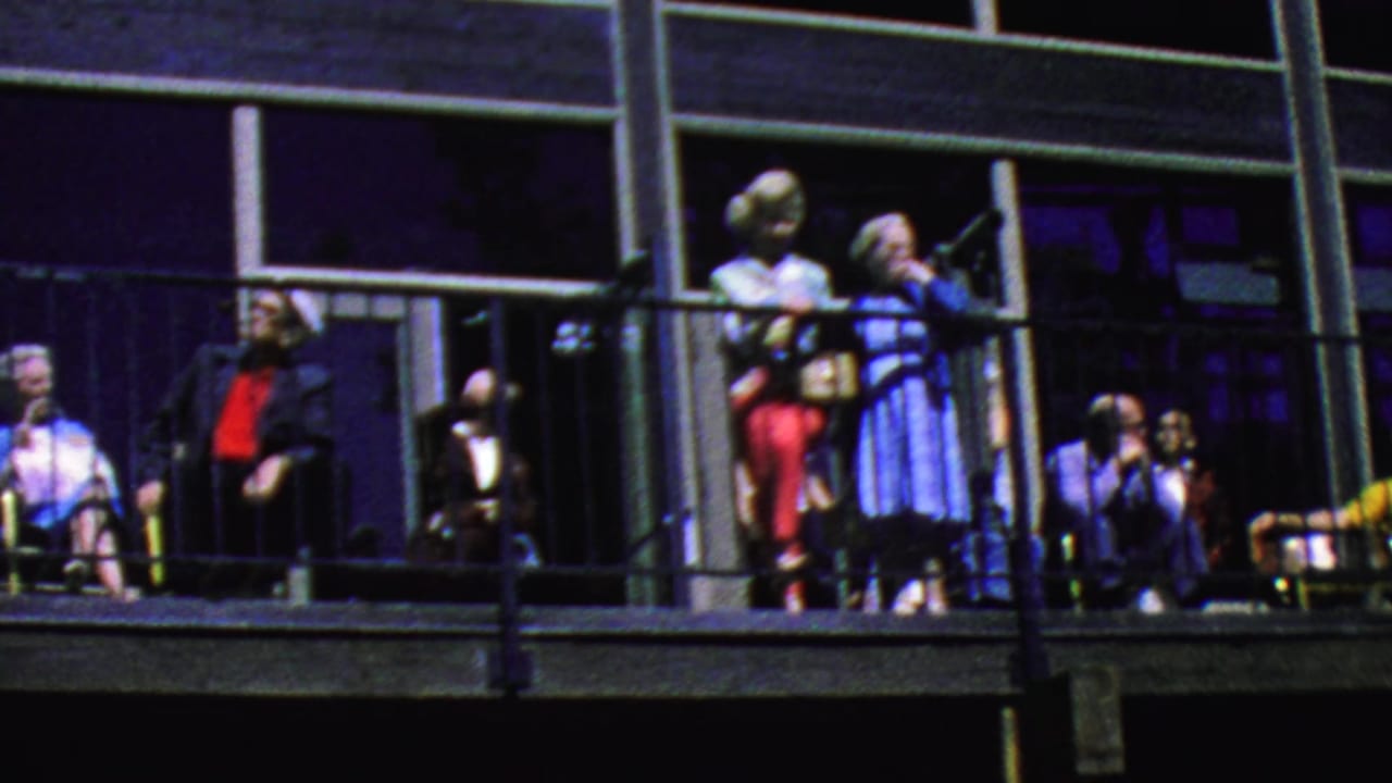 Adults stand and sit on a balcony behind black metal railings and large dark windows.
