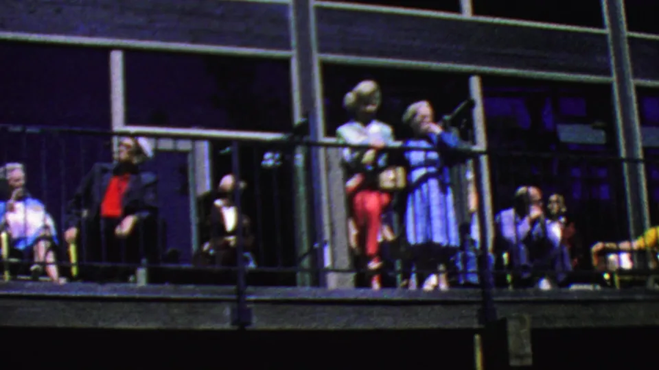 Adults at the Palm Springs Aerial Tramway in 1964 appear on an exterior balcony, with seated spectators, standing figures, black railings, and large window panels behind them.