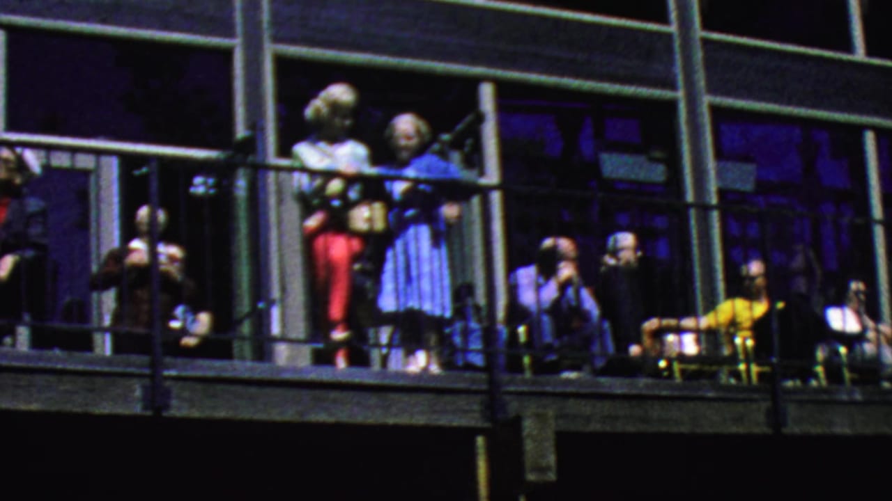 People gather along a balcony railing, with chairs and large windows behind them.