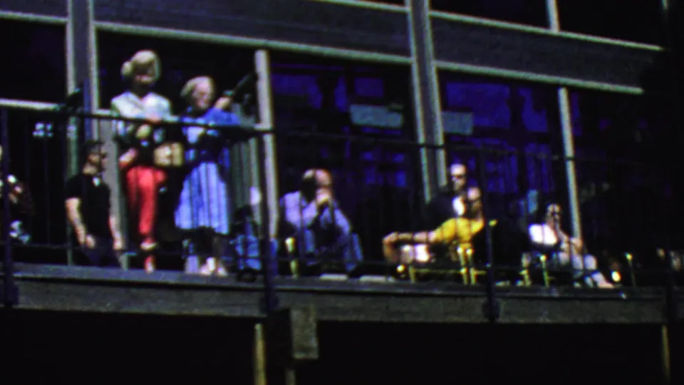 Adults on the Palm Springs tramway balcony stand and sit behind black railings, with a blue-dressed woman near center and mid-century chairs visible along the deck.
