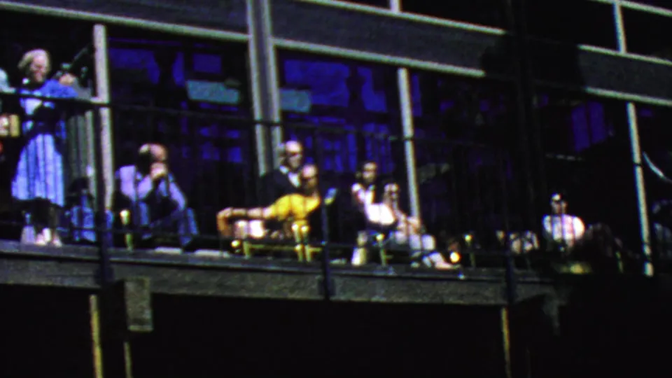 The final 1964 Palm Springs frame shows visitors spread across the balcony, with a standing woman near the railing, seated adults, yellow clothing, chairs, and dark window panels.