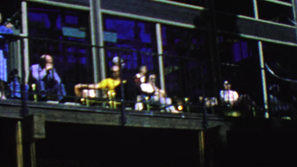 Adults relax on a long balcony at the Palm Springs Aerial Tramway in 1964, with patio chairs, railings, and blue-tinted window reflections visible.
