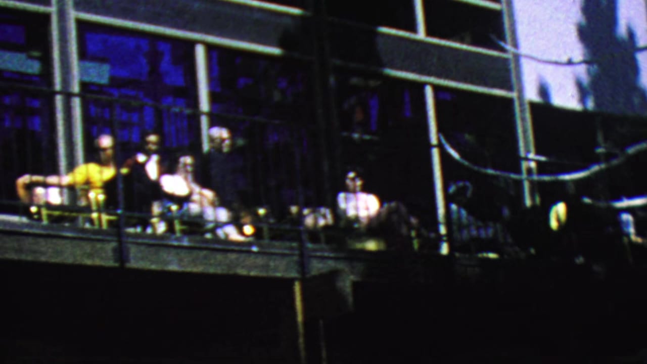Several adults sit in chairs on a balcony with glass windows behind them.