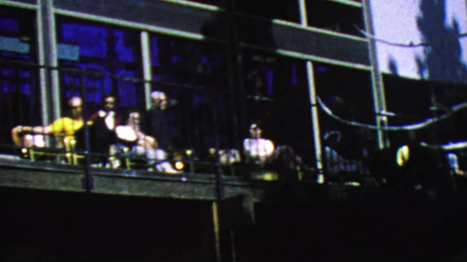 A group of visitors sits and talks on the Palm Springs Aerial Tramway balcony in 1964, framed by railings, chairs, and dark blue window reflections.