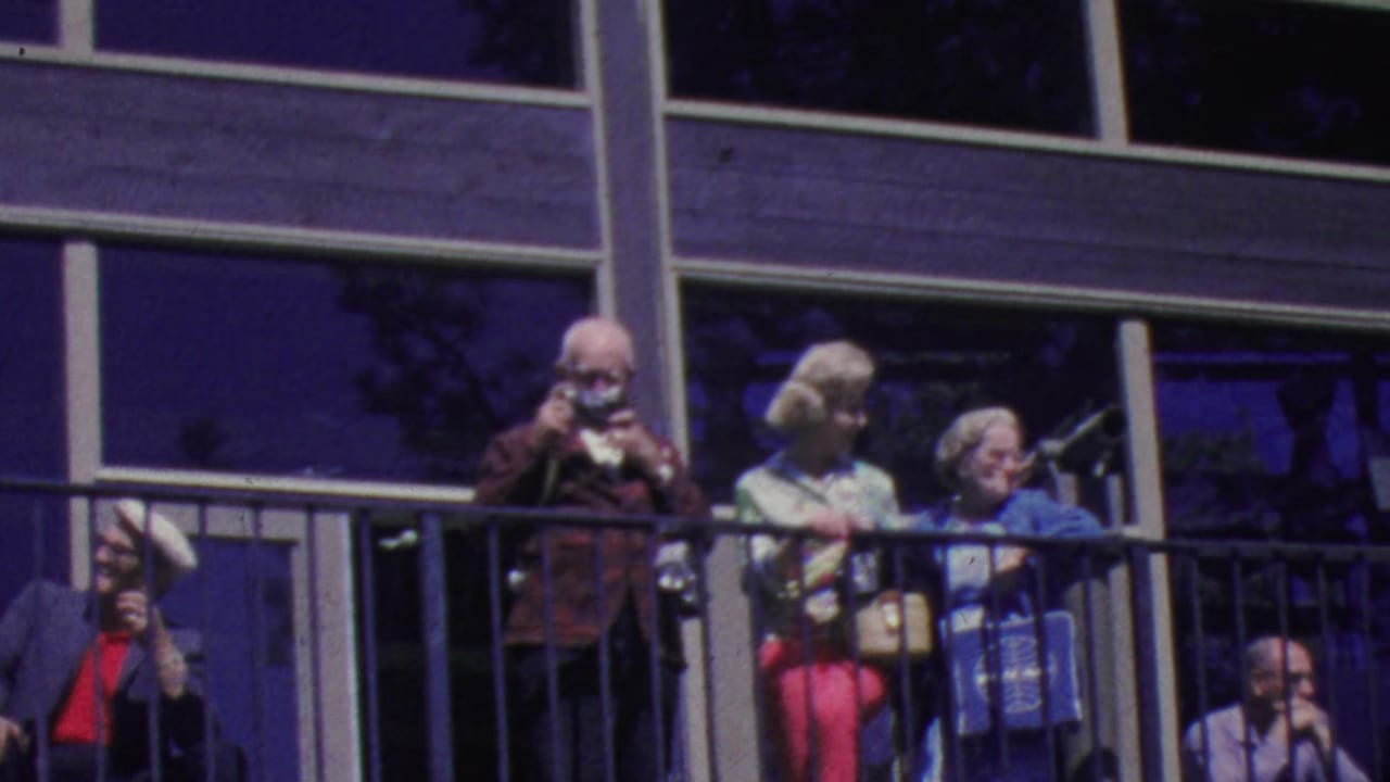 Several older adults stand on a balcony as one man photographs the view.