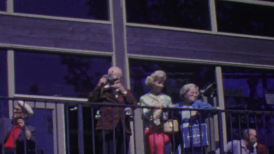 In this 1964 Palm Springs tramway frame, a man with a camera stands near two women in bright coats behind the exterior balcony railing.