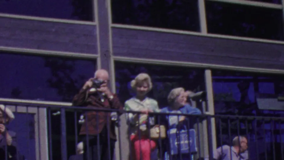 Older tourists at the Palm Springs Aerial Tramway in 1964 remain gathered along a black railing outside a glass-fronted building, with cameras and bright coats visible.