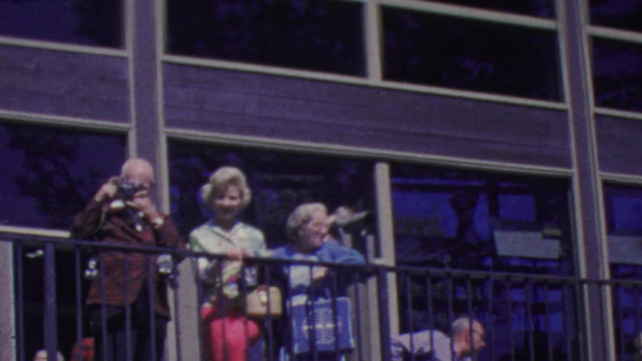1964 Palm Springs Aerial Tramway spectators, including older adults and a person using a small camera, gather behind a balcony railing outside a modern windowed building.