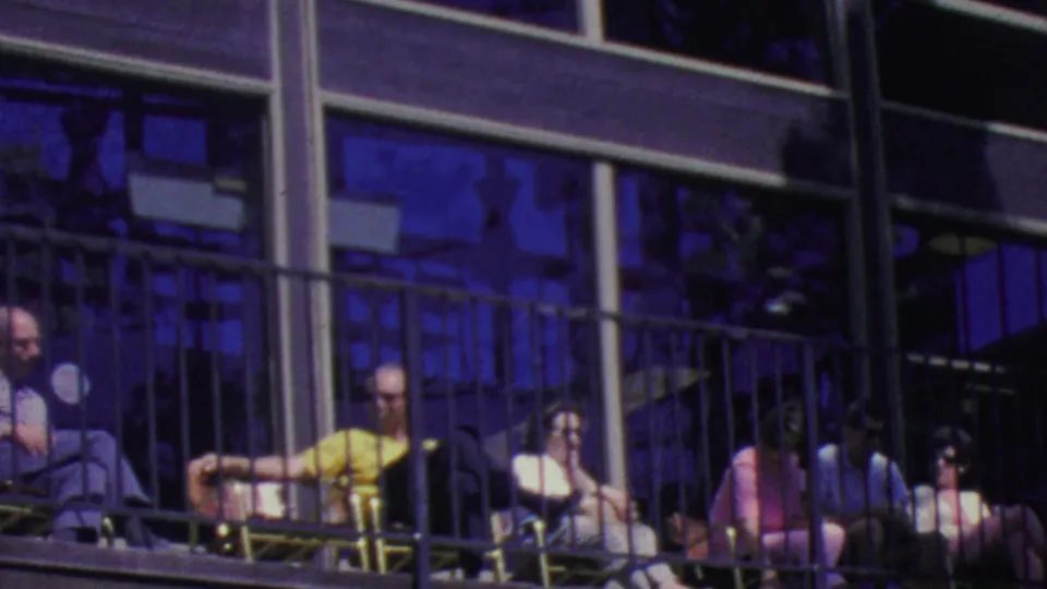 A row of 1960s spectators sits on a Palm Springs balcony behind a dark railing, including a man in a bright yellow shirt and several people in sunglasses.