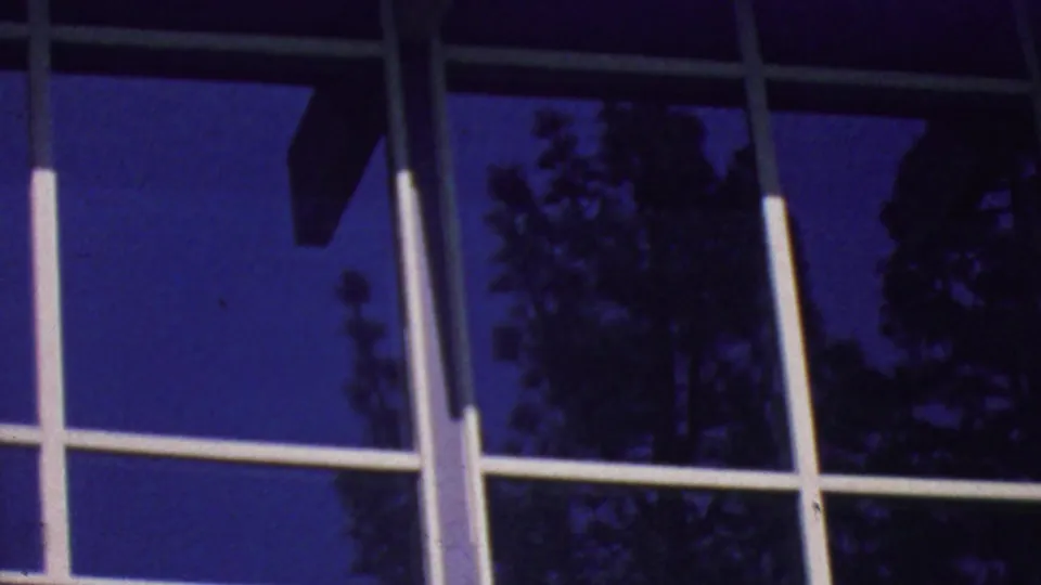 Palm Springs 1964 home-movie still of an angled modernist facade with reflective windows, white rails, and silhouetted trees visible in the glass.