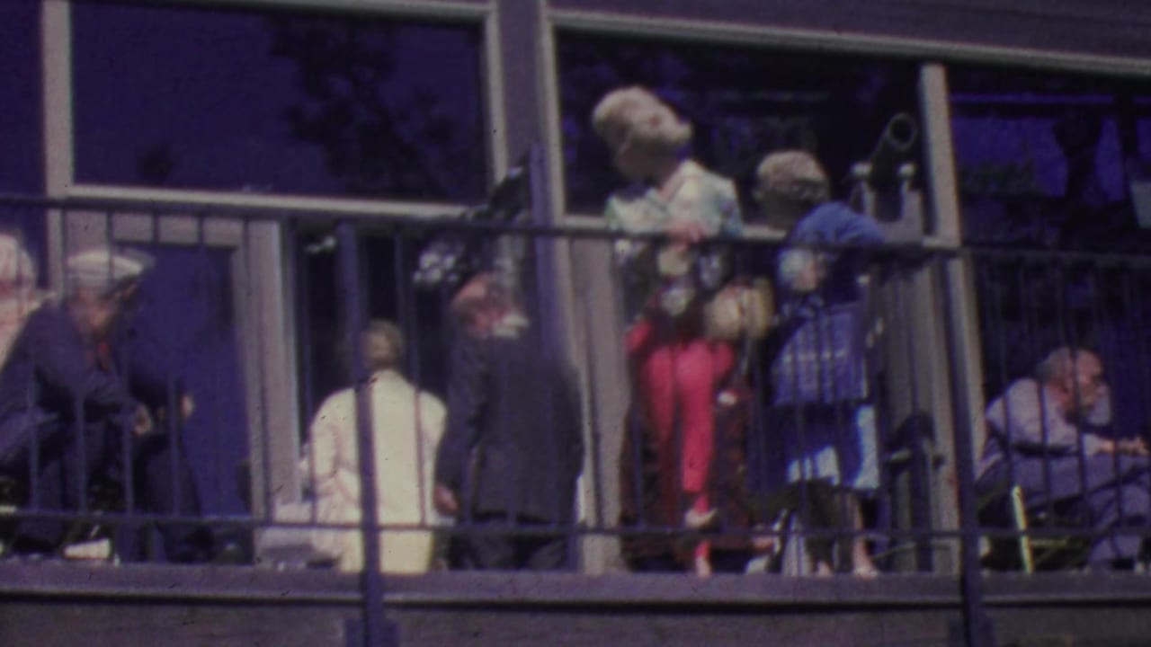 Several adults stand and sit along a balcony with railings and reflective windows behind them.