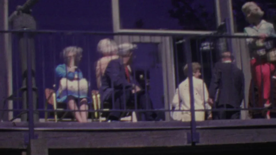 1964 home-movie frame from the Palm Springs Aerial Tramway showing adults in mid-century clothing gathered on a balcony, with chairs, railings, and reflective windows visible.