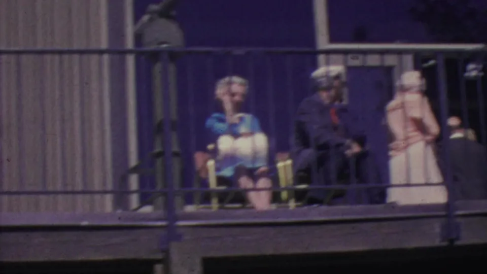 1964 Palm Springs Aerial Tramway frame showing a seated woman in a blue top, adults nearby, a light-colored dress, balcony railings, and the building's large windows.