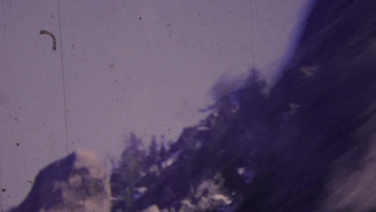 1964 Palm Springs-area 8mm frame showing a blurred rocky mountain slope, dark trees, and pale sky from a moving aerial tramway viewpoint.