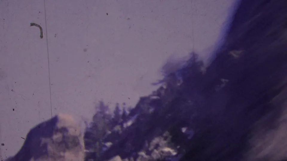 1964 Palm Springs-area 8mm frame showing a blurred rocky mountain slope, dark trees, and pale sky from a moving aerial tramway viewpoint.