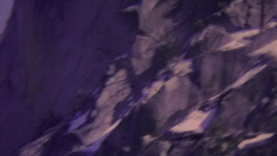1964 Palm Springs-area mountain view from an aerial tramway, with rocky slopes, snow traces, and trees rendered in soft purple-shifted 8mm color.