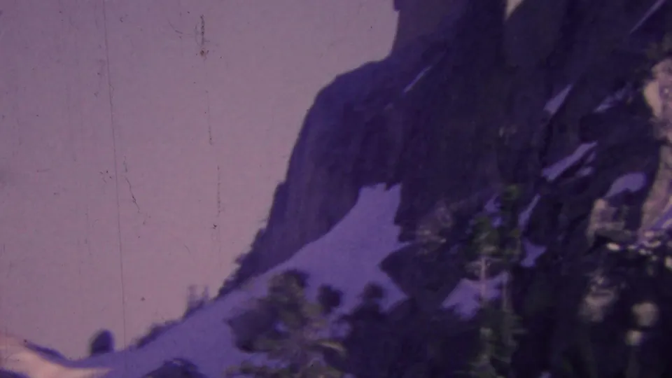 1964 8mm Palm Springs aerial tramway view of a snow-streaked mountain ridge, dark rock formations, and evergreen trees below the tramway perspective.