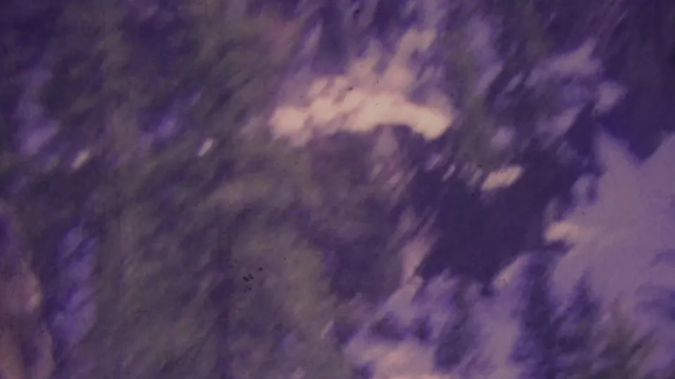 1964 Palm Springs aerial tramway frame showing a snowy, tree-lined mountain slope and dark rocky terrain, softened by handheld movement and aged-film color shift.