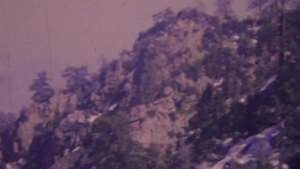 A soft 1964 8mm view across Palm Springs-area rocky mountain terrain, with dark trees silhouetted against a muted sky.