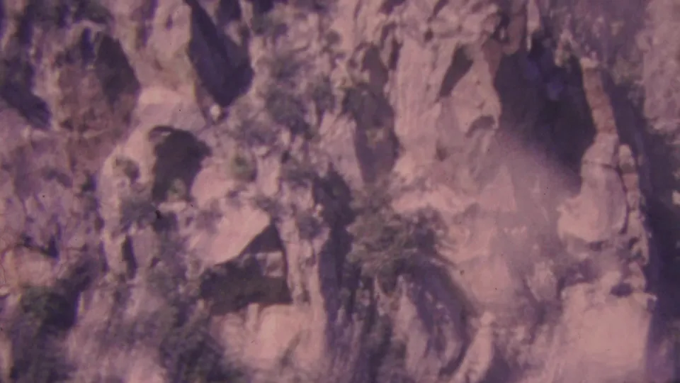 Palm Springs-area 1964 8mm frame showing a rugged granite cliff face with ledge vegetation, pine trees, and dark crevices in the rock.