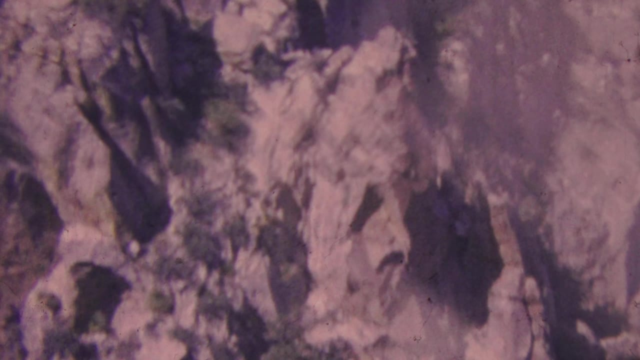 1964 8mm Palm Springs area view of a rugged cliff face with jagged stone, sparse shrubs, and deep shadowed recesses.