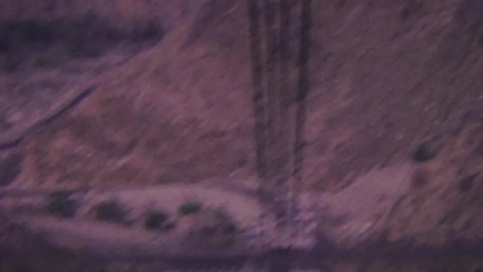 1964 Palm Springs 8mm frame showing a rugged desert mountain slope crossed by aerial tramway cables, with sparse shrubs and soft distant terrain.