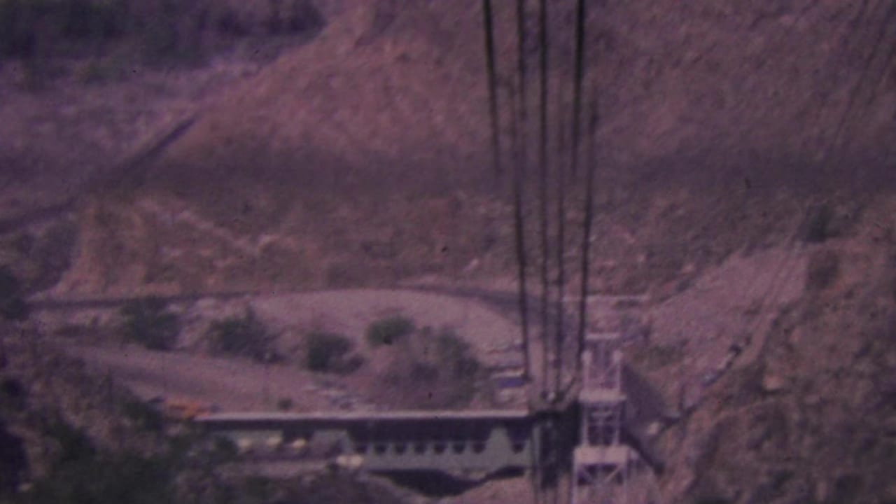 1964 Palm Springs 8mm frame showing the aerial tramway support area and cable lines set into a steep, sparsely vegetated mountain slope.