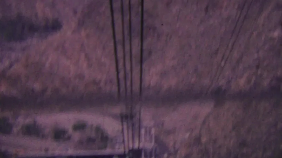 1964 Palm Springs 8mm frame with dark tramway cables in the foreground over a blurred arid hillside, sparse shrubs, and faint road or path forms below.