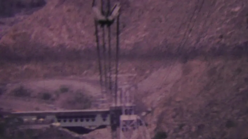 1964 Palm Springs 8mm frame showing a tramway support tower and cable car or station structure beside a steep rocky desert slope.