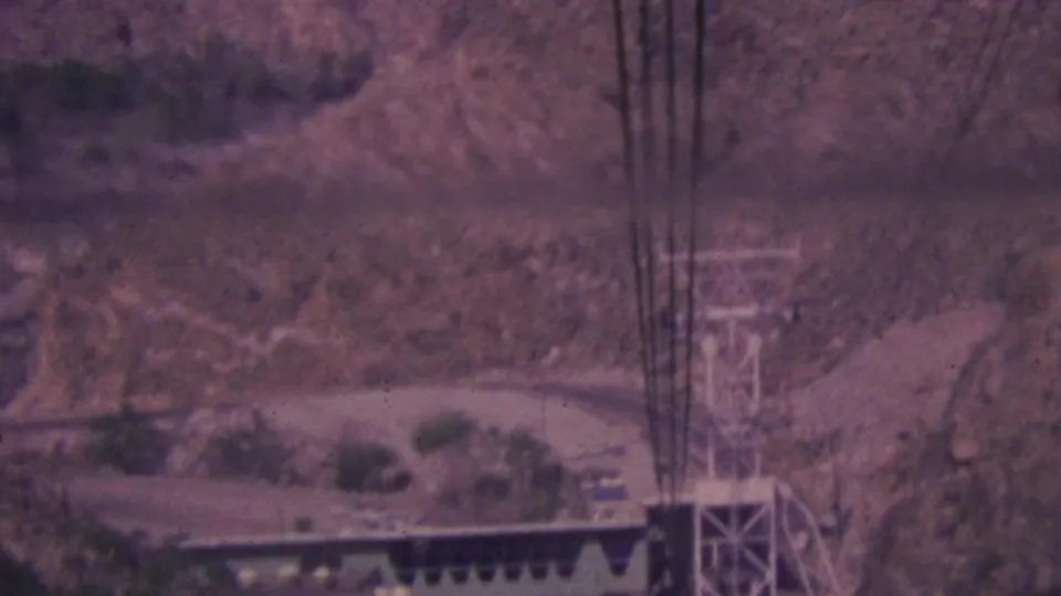 1964 Palm Springs 8mm view of a metal transmission tower and vertical lines near a winding road on a rugged desert mountainside.