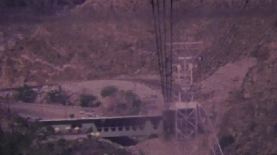 1964 Palm Springs aerial tramway structures appear across a rugged desert canyon, with towers, cables, and station-like platforms seen through soft 8mm film grain.
