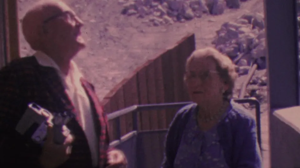 An older couple at the 1964 Palm Springs aerial tramway area look upward near a railing, with the man holding a camera and rocky desert terrain behind them.