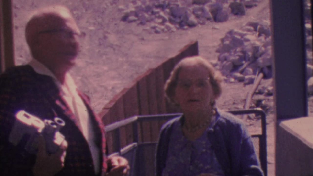 An older couple in 1964 Palm Springs tramway home-movie footage sit by the window, with the man in glasses and a plaid jacket and the woman in blue facing the camera.