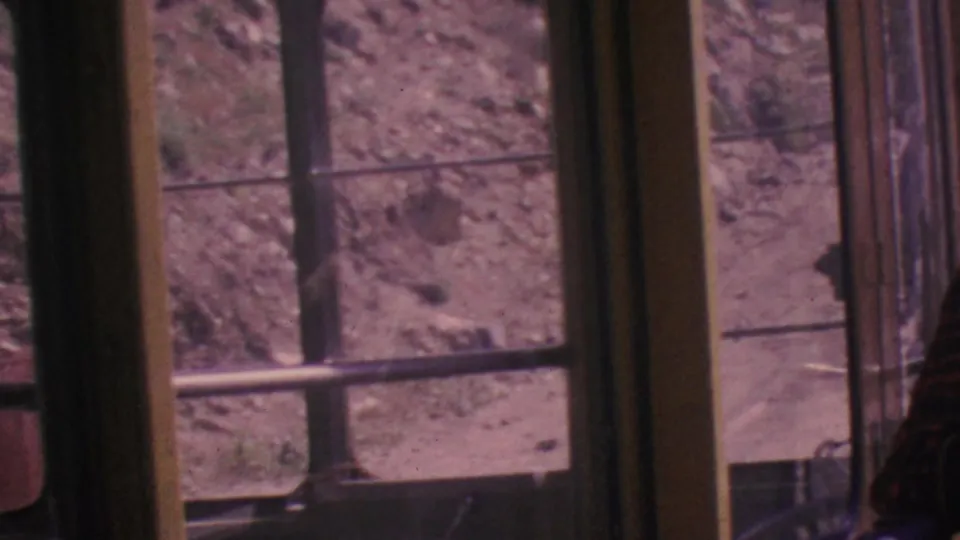 The final frame looks across the tram-car windows and metal railings toward the rocky arid hillside, with only a partial passenger edge visible at the side.