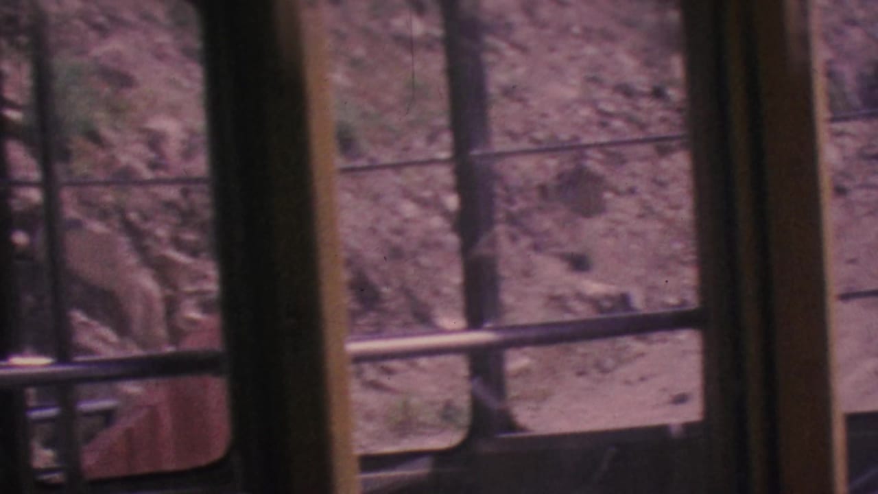 Inside the Palm Springs Aerial Tramway in 1964, older passengers stand by tall window frames as rocky sandy ground passes outside.