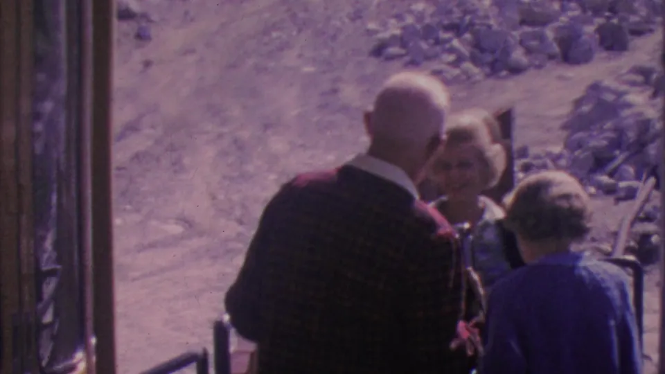 The older trio stands close together near the tram entrance, appearing to converse while the desert slope and metal railing remain visible outside.