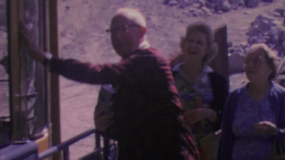 The older man in a plaid jacket gestures beside smiling women inside the Palm Springs tram, with the sandy rocky landscape visible through the open side.