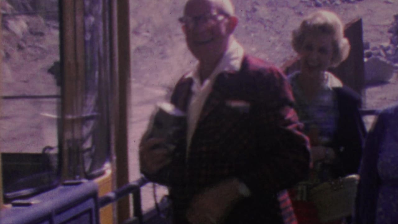Older man with glasses holds a camera while smiling near a tram doorway.