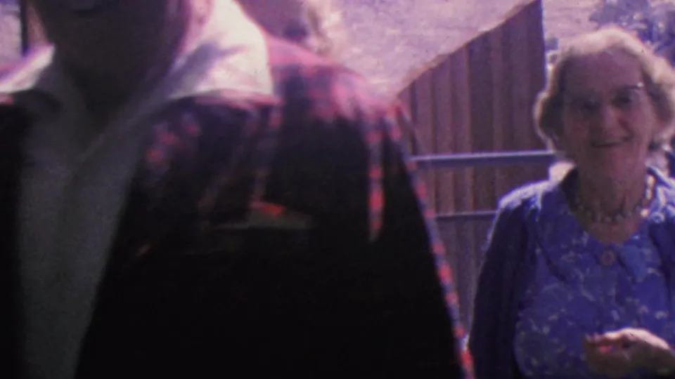 An older woman in a blue dress smiles toward the camera near the tram doorway as the man in the patterned jacket passes through the foreground.